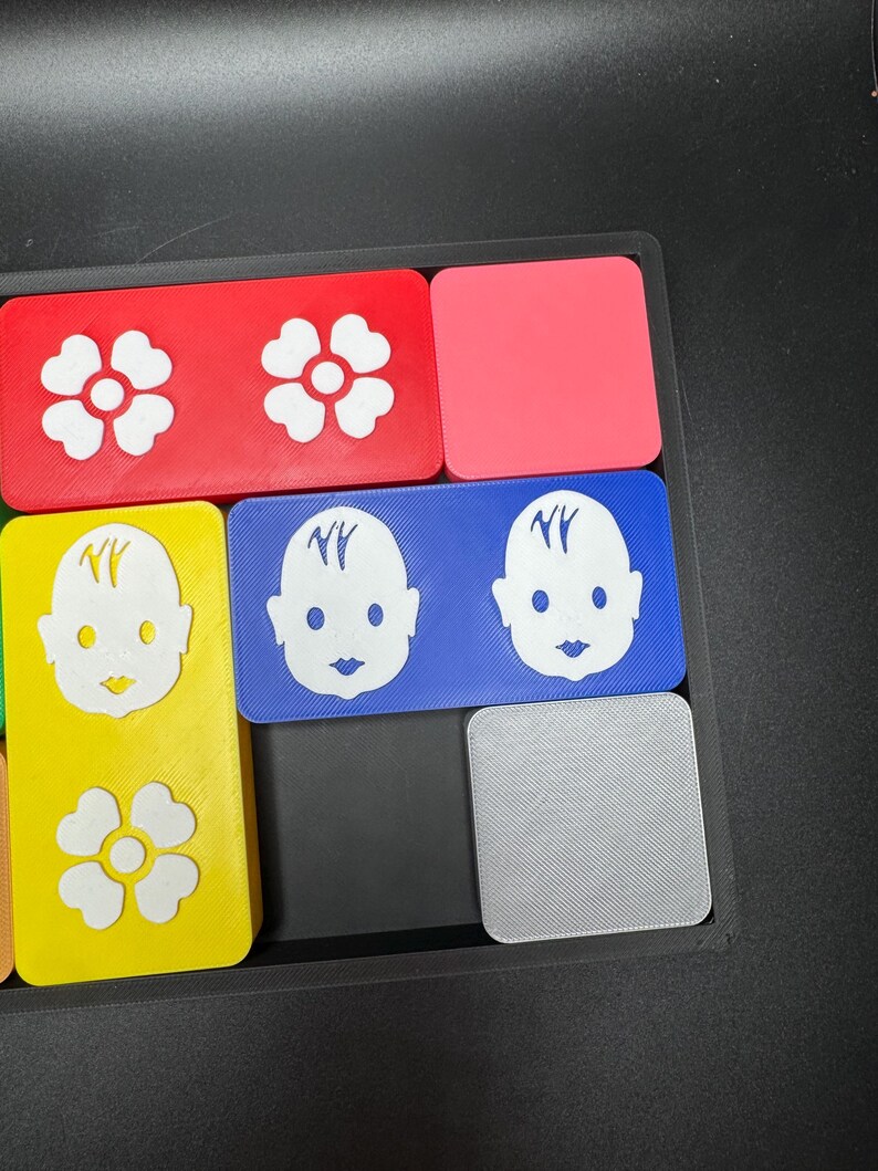 May include: A set of colorful square tiles with different designs. The tiles feature a white flower, a baby face with the letters "NY" on the forehead, and a gray tile with a textured surface.