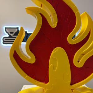 May include: A three-dimensional puzzle in the shape of a flame. The puzzle is primarily yellow with a red interior, and is displayed on a white surface. The flame design is composed of interlocking pieces, suggesting a jigsaw puzzle.