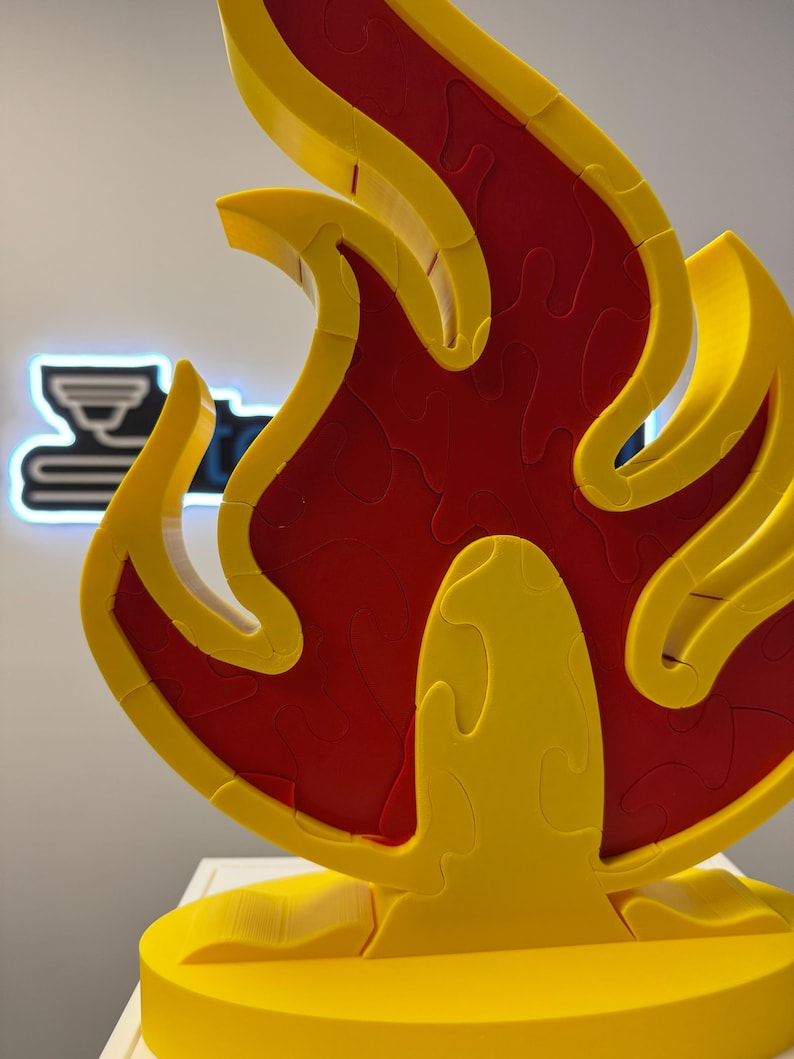 May include: A three-dimensional puzzle in the shape of a flame. The puzzle is primarily yellow with a red interior, and is displayed on a white surface. The flame design is composed of interlocking pieces, suggesting a jigsaw puzzle.