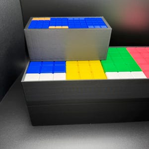 May include: A set of three black boxes with colorful square tiles inside. The top box has blue tiles with a few orange tiles. The middle box has blue, yellow, white, and green tiles. The bottom box has green, white, and red tiles.