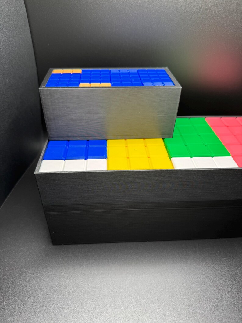 May include: A set of three black boxes with colorful square tiles inside. The top box has blue tiles with a few orange tiles. The middle box has blue, yellow, white, and green tiles. The bottom box has green, white, and red tiles.