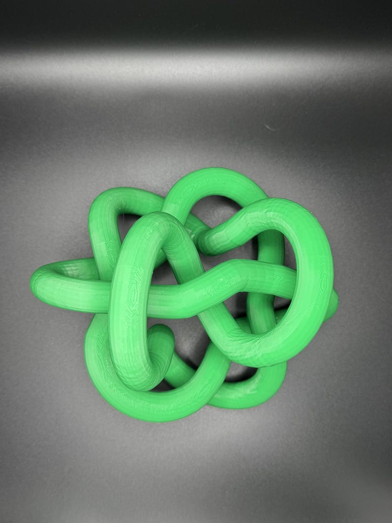 K&ouml;nnte beinhalten: Eine gr&uuml;ne Plastikknotenskulptur. Der Knoten besteht aus einem einzigen St&uuml;ck Plastik, das verdreht und gedreht wird, um ein komplexes Design zu schaffen.