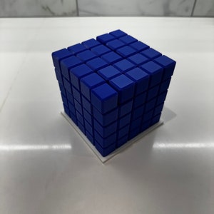 May include: A blue cube made of smaller cubes, each with a slightly textured surface. The cube is sitting on a white base.