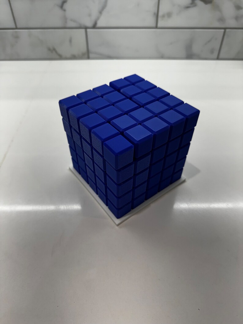 May include: A blue cube made of smaller cubes, each with a slightly textured surface. The cube is sitting on a white base.