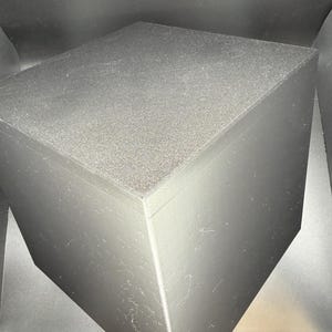 May include: A solid black cube with a matte finish and visible linear texture lines on its vertical sides. The top surface has a slightly granular, textured appearance, while the overall structure maintains sharp, clean edges and a geometric form.