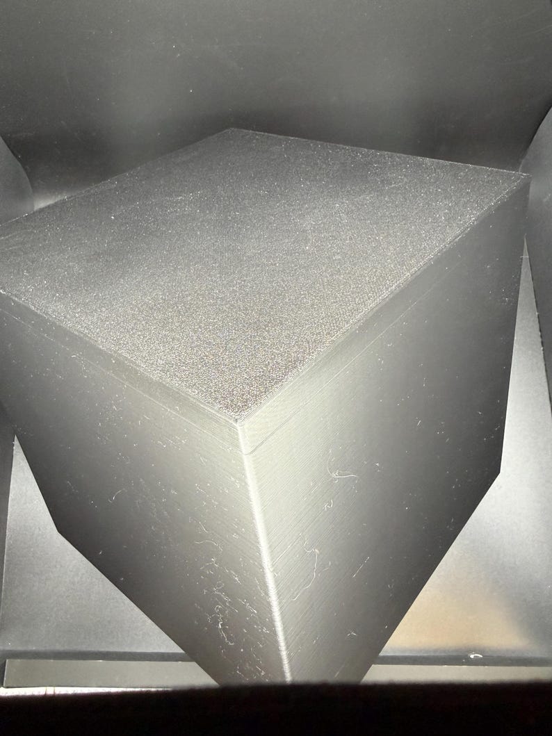 May include: A solid black cube with a matte finish and visible linear texture lines on its vertical sides. The top surface has a slightly granular, textured appearance, while the overall structure maintains sharp, clean edges and a geometric form.