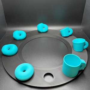 Op de afbeelding: Een verzameling turquoisekleurige minidonuts en mokken gerangschikt op een zwarte cirkelvormige basis. De donuts hebben een gat in het midden en de mokken hebben oren. De objecten zijn gelijkmatig verdeeld over de basis.