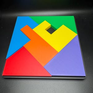 May include: A colorful wooden puzzle with six pieces, including a blue, green, yellow, orange, red, and purple piece. The pieces are arranged to form a square shape with a small hole in the center.