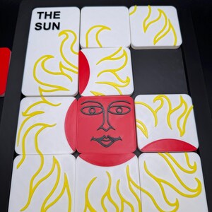 May include: A set of white square tiles with yellow flame designs and a red sun face. One tile reads "THE SUN" in black text. The tiles are arranged on a black surface, creating a puzzle or game.