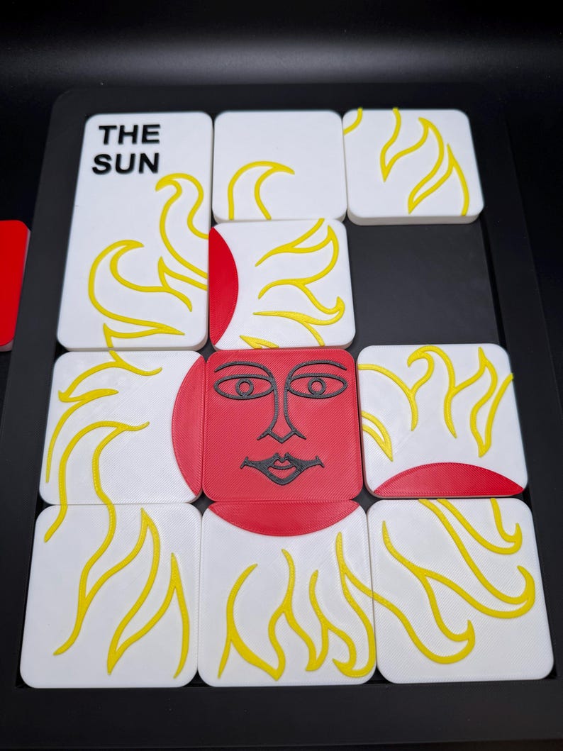 May include: A set of white square tiles with yellow flame designs and a red sun face. One tile reads "THE SUN" in black text. The tiles are arranged on a black surface, creating a puzzle or game.