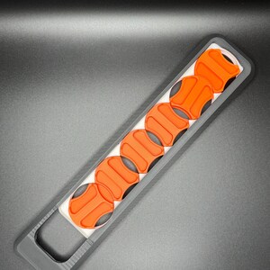 May include: A black plastic holder with a row of six orange round buttons. The holder has a handle on one end.