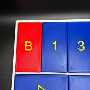 May include: A close-up of a red and blue game board with yellow letters and numbers. The board has a white frame and the letters and numbers are raised. The letters are "B" and the numbers are "1", "3", and "4".