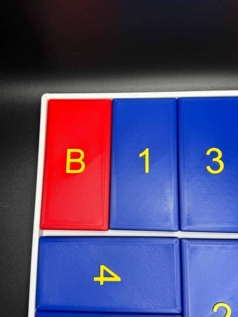 May include: A close-up of a red and blue game board with yellow letters and numbers. The board has a white frame and the letters and numbers are raised. The letters are "B" and the numbers are "1", "3", and "4".