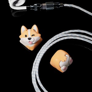 Op de afbeelding: Een close-up van een oplaadkabel met een wit en grijs gevlochten ontwerp. De kabel heeft een magnetische connector en twee corgi-vormige bedels, een knipoogt en steekt zijn tong uit, de andere toont zijn achterwerk.
