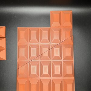 Op de afbeelding: Een grote, oranje-bruine chocolade reep met een getextureerd oppervlak, gebroken in meerdere stukken. De reep is gemaakt van een raster van vierkanten.