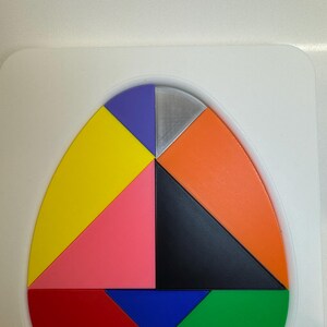 Op de afbeelding: Een kleurrijke, eivormige puzzel met acht stukken. De stukken zijn in de volgende kleuren: geel, roze, zwart, oranje, blauw, groen, paars en zilver.