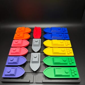 Op de afbeelding: Een set van 10 kleurrijke speelgoedbootjes, gerangschikt in een rasterpatroon op een zwart oppervlak. De bootjes zijn gemaakt van plastic en hebben verschillende kleuren, waaronder rood, oranje, geel, groen, blauw en paars. Er is ook een grijs bootje in het midden van het raster.