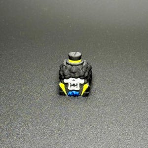 May include: A black and yellow 3D printed figurine of a skeleton wearing a top hat and bow tie. The figurine has a skull face with white eyes and a black afro.