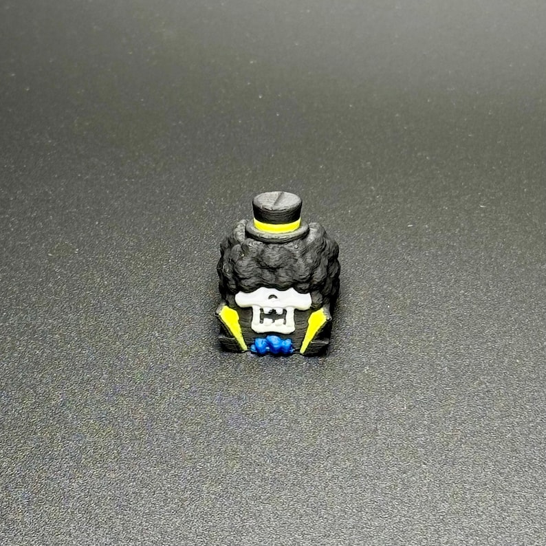 May include: A black and yellow 3D printed figurine of a skeleton wearing a top hat and bow tie. The figurine has a skull face with white eyes and a black afro.