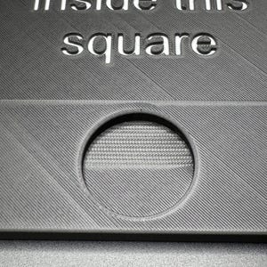 K&ouml;nnte beinhalten: Ein graues, strukturiertes Quadrat mit dem ausgeschnittenen Text "10 Kreise in diesem Quadrat". Ein kreisf&ouml;rmiges Loch ist im unteren Teil des Quadrats sichtbar.