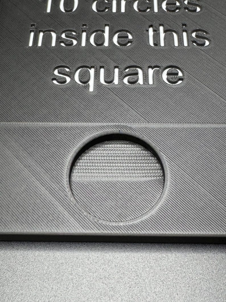 K&ouml;nnte beinhalten: Ein graues, strukturiertes Quadrat mit dem ausgeschnittenen Text "10 Kreise in diesem Quadrat". Ein kreisf&ouml;rmiges Loch ist im unteren Teil des Quadrats sichtbar.