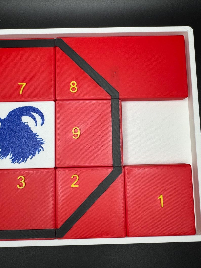 May include: A red puzzle with black trim and yellow numbers 1 through 9. The puzzle is arranged in a grid pattern with a goat illustration in the center.