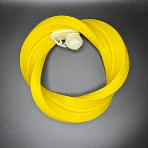 Może przedstawiać: Jasnoż&oacute;łty silikonowy pierścień z białą plastikową nakładką. Pierścień jest skręcony w kształt &oacute;semki.