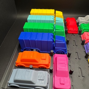 May include: A selection of brightly coloured toy trucks and cars in shades of blue, green, yellow, orange, red, and pink. The vehicles are arranged in a black tray, showcasing a variety of vehicle types.
