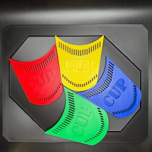 May include: Four colorful, plastic cup holders in red, yellow, blue, and green, each with the word "CUP" embossed. The holders are arranged on a black, geometric tray.