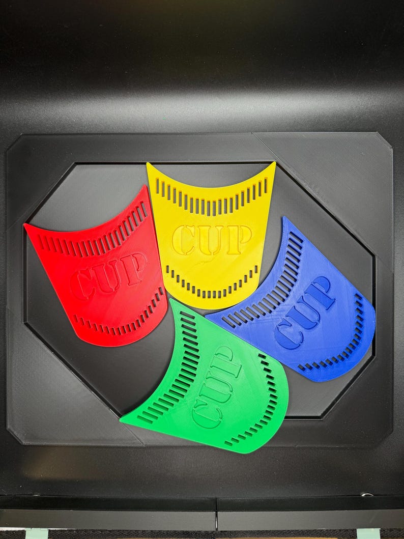 May include: Four colorful, plastic cup holders in red, yellow, blue, and green, each with the word "CUP" embossed. The holders are arranged on a black, geometric tray.