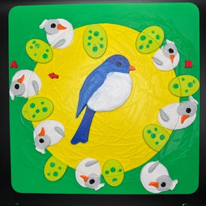 May include: A colorful, handcrafted bird-themed puzzle. The puzzle features a blue and white bird in the center, surrounded by white bird figures and green eggs on a yellow background, all set on a green square.