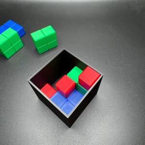 May include: A black square container holds a puzzle with red, green, and blue blocks. Several loose blocks of green and blue are scattered nearby. The blocks are arranged to fit within the container.