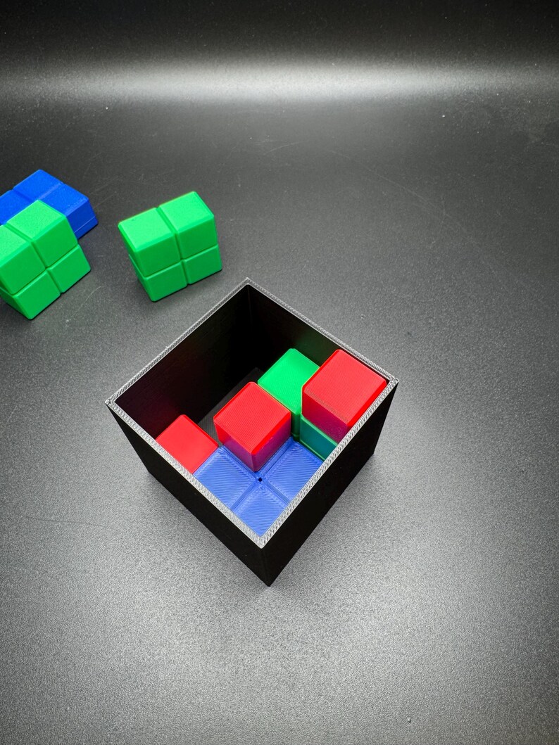 May include: A black square container holds a puzzle with red, green, and blue blocks. Several loose blocks of green and blue are scattered nearby. The blocks are arranged to fit within the container.