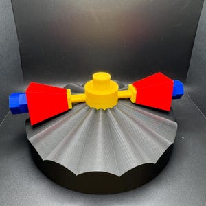 May include: A 3D-printed, multi-colored object with a fan-like base. The base is black, with a gray, ridged top. A yellow central component extends to red and blue geometric shapes. The object is on a black surface.