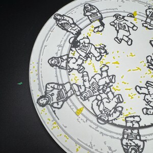 Puede incluir: Plato de cer&aacute;mica blanco con un dise&ntilde;o de astronauta negro y amarillo. El plato presenta m&uacute;ltiples figuras de astronautas en un patr&oacute;n circular, con motas amarillas y un borde gris. El dise&ntilde;o es una representaci&oacute;n art&iacute;stica y l&uacute;dica de la exploraci&oacute;n espacial.