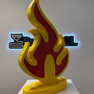 May include: A yellow and red flame-shaped sculpture. The sculpture has a yellow outline and a red interior with a textured pattern. The flame is mounted on a yellow base. The background features a neon sign with the text "I+CL".