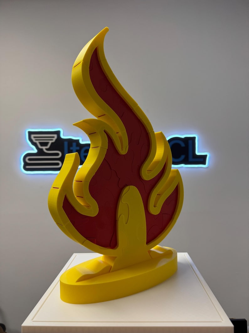 May include: A yellow and red flame-shaped sculpture. The sculpture has a yellow outline and a red interior with a textured pattern. The flame is mounted on a yellow base. The background features a neon sign with the text "I+CL".