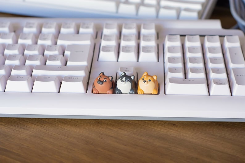 May include: A white mechanical keyboard with three custom keycaps shaped like cartoon dogs. The keycaps are brown, black and white, and tan and cream colored. The keyboard sits on a wooden surface.