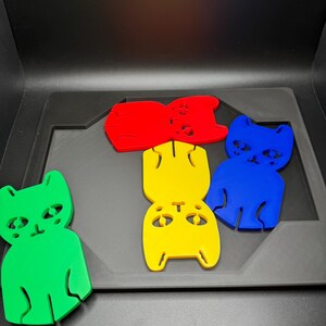 May include: A collection of colorful cat-shaped cutouts in green, red, yellow, and blue. The cat figures have simple, stylized features and are arranged on a dark gray surface. The cutouts appear to be made of a solid material.