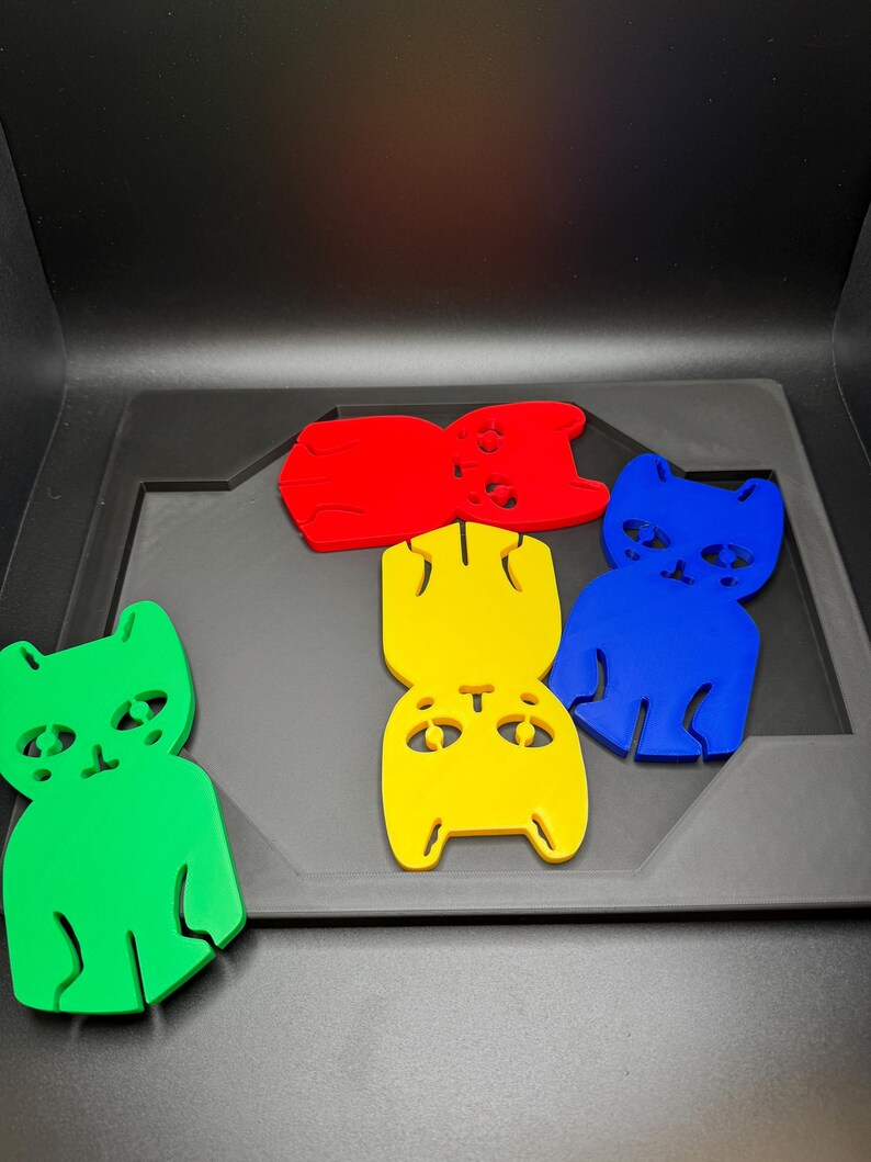 May include: A collection of colorful cat-shaped cutouts in green, red, yellow, and blue. The cat figures have simple, stylized features and are arranged on a dark gray surface. The cutouts appear to be made of a solid material.