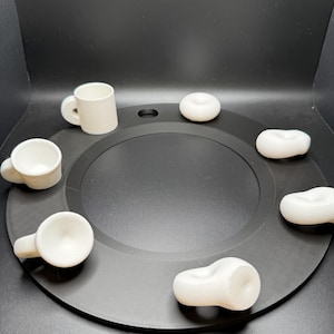 Op de afbeelding: Een verzameling kleine, witte keramische mokken en donutvormige objecten gerangschikt op een zwarte ronde tray. De mokken hebben handvatten en afgeronde lichamen. De donuts zijn glad en solide. De objecten zijn gelijkmatig verdeeld over de tray.
