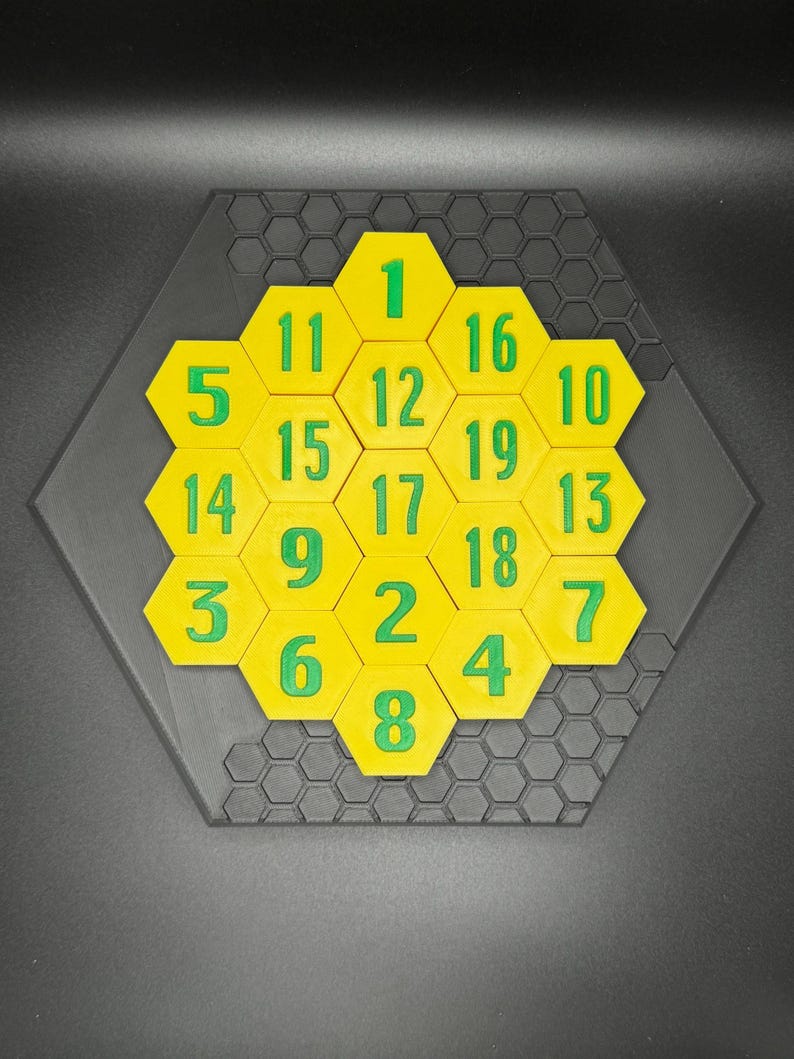 Puede incluir: Un rompecabezas hexagonal amarillo con n&uacute;meros verdes del 1 al 19 en cada pieza. El rompecabezas est&aacute; sobre una base hexagonal negra.