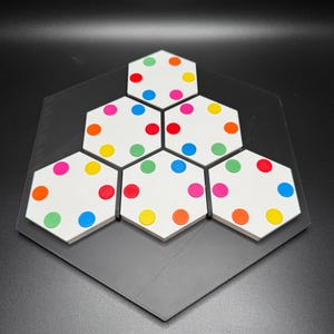 May include: A hexagonal game board with six white hexagonal tiles, each with six colored dots. The dots are red, orange, yellow, green, blue, and pink.