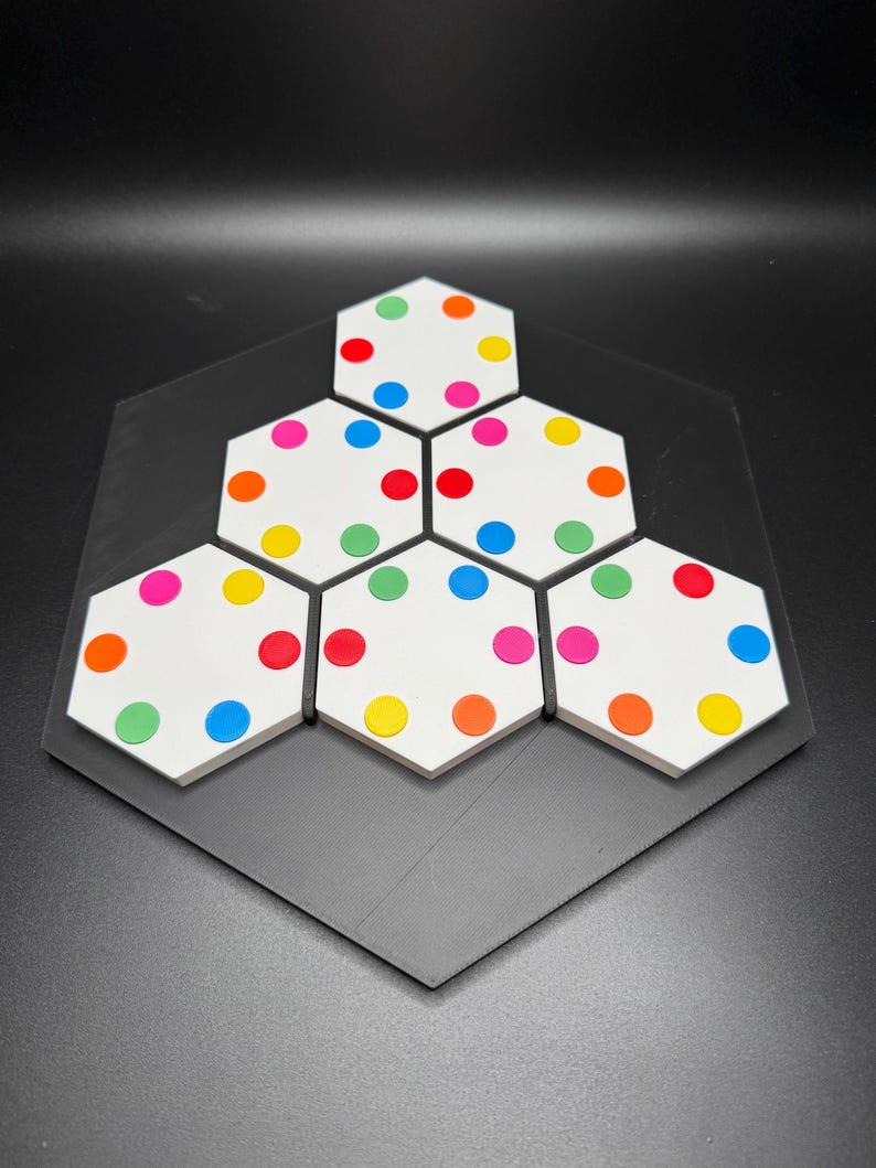 May include: A hexagonal game board with six white hexagonal tiles, each with six colored dots. The dots are red, orange, yellow, green, blue, and pink.