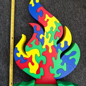 Puede incluir: Un rompecabezas de madera multicolor con forma de llama sobre una base verde, colocado junto a una cinta m&eacute;trica amarilla que muestra una altura de aproximadamente 63,5 cm. El rompecabezas consta de piezas entrelazadas en rojo, amarillo, azul y verde.