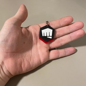May include: A black, red, and white hexagonal keychain with a fist graphic. The keychain is held in a hand. The fist graphic is white and the bottom layer of the keychain is red.