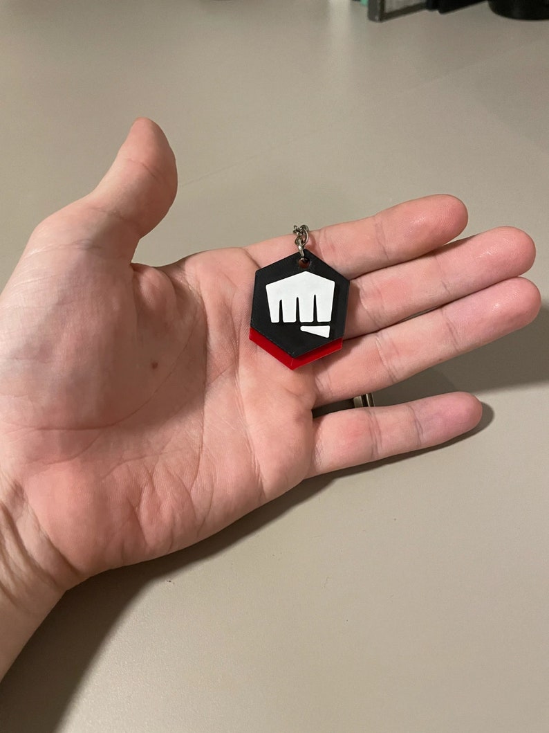 May include: A black, red, and white hexagonal keychain with a fist graphic. The keychain is held in a hand. The fist graphic is white and the bottom layer of the keychain is red.