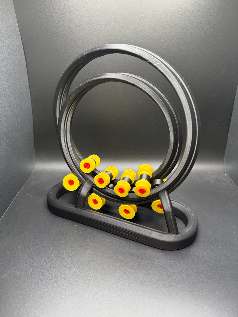 May include: A black, 3D-printed abacus-style toy with a double-loop track. The toy features yellow and red beads that move along the track. The base is also black, and the background is a dark gray.