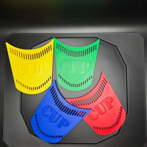 May include: Four colorful, plastic cup-shaped objects in yellow, green, blue, and red, each with the word "CUP" printed on them. The objects are arranged on a black surface.