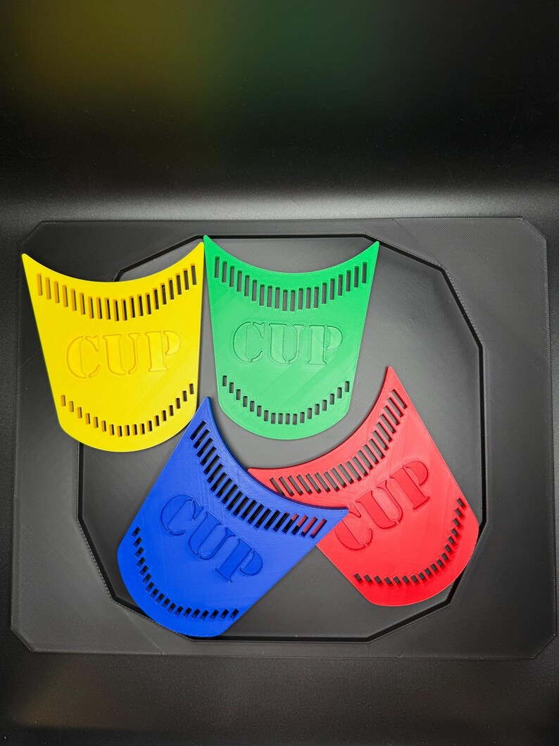 May include: Four colorful, plastic cup-shaped objects in yellow, green, blue, and red, each with the word "CUP" printed on them. The objects are arranged on a black surface.